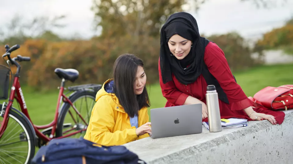Foto. Två studenter utomhus tittar på dator. I bakgrunden en cykel. Fotograf: Johan Persson